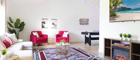 Un salon avec un ventilateur de plafond, une cheminée, un grand tableau représentant une plage et un fauteuil rouge.