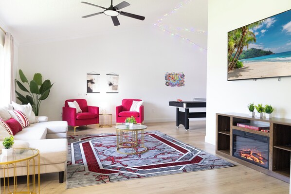 A living room with a ceiling fan, a fireplace, a large painting of a beach, and a red armchair.