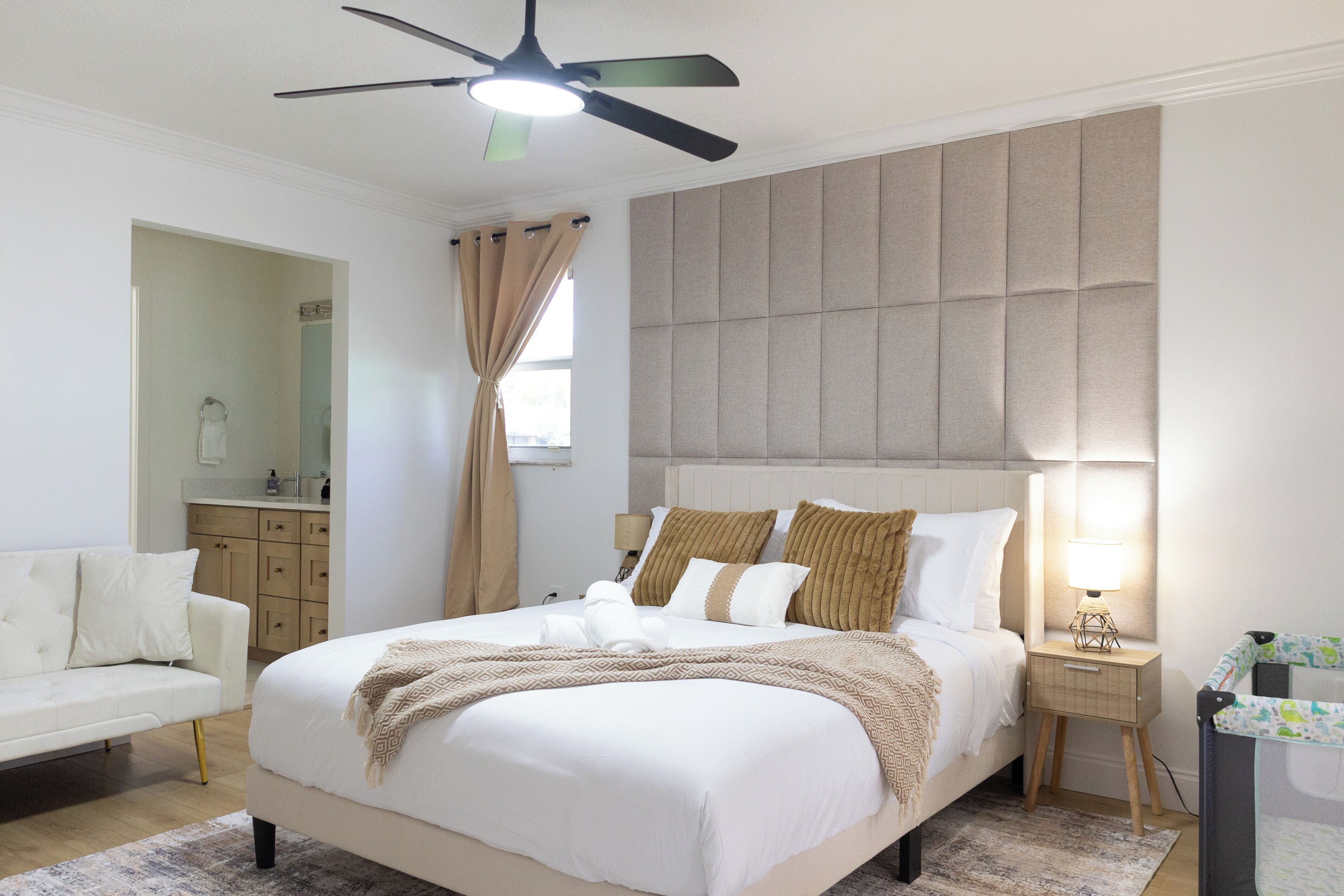 A bedroom with a bed, a nightstand, a lamp, a ceiling fan, and a view of a bathroom.