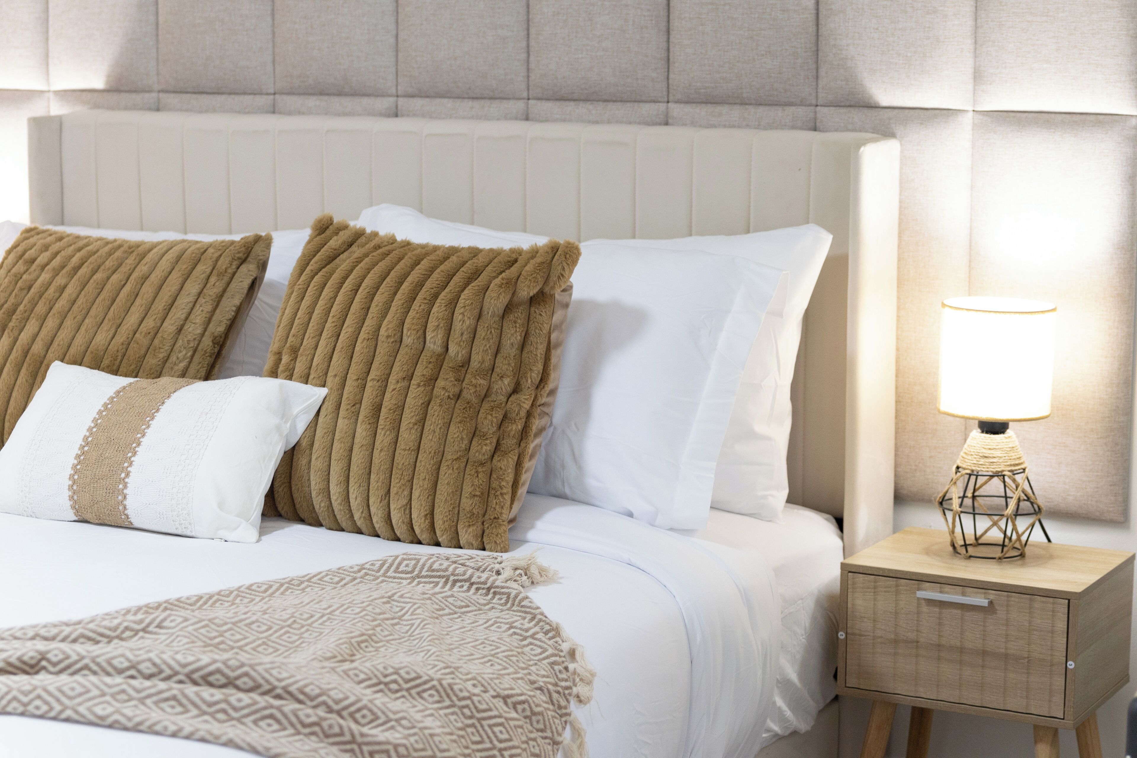 Een opgemaakt bed met een patroonkleed, een nachtkastje met een lamp en een hoofdbord met een structuur.
