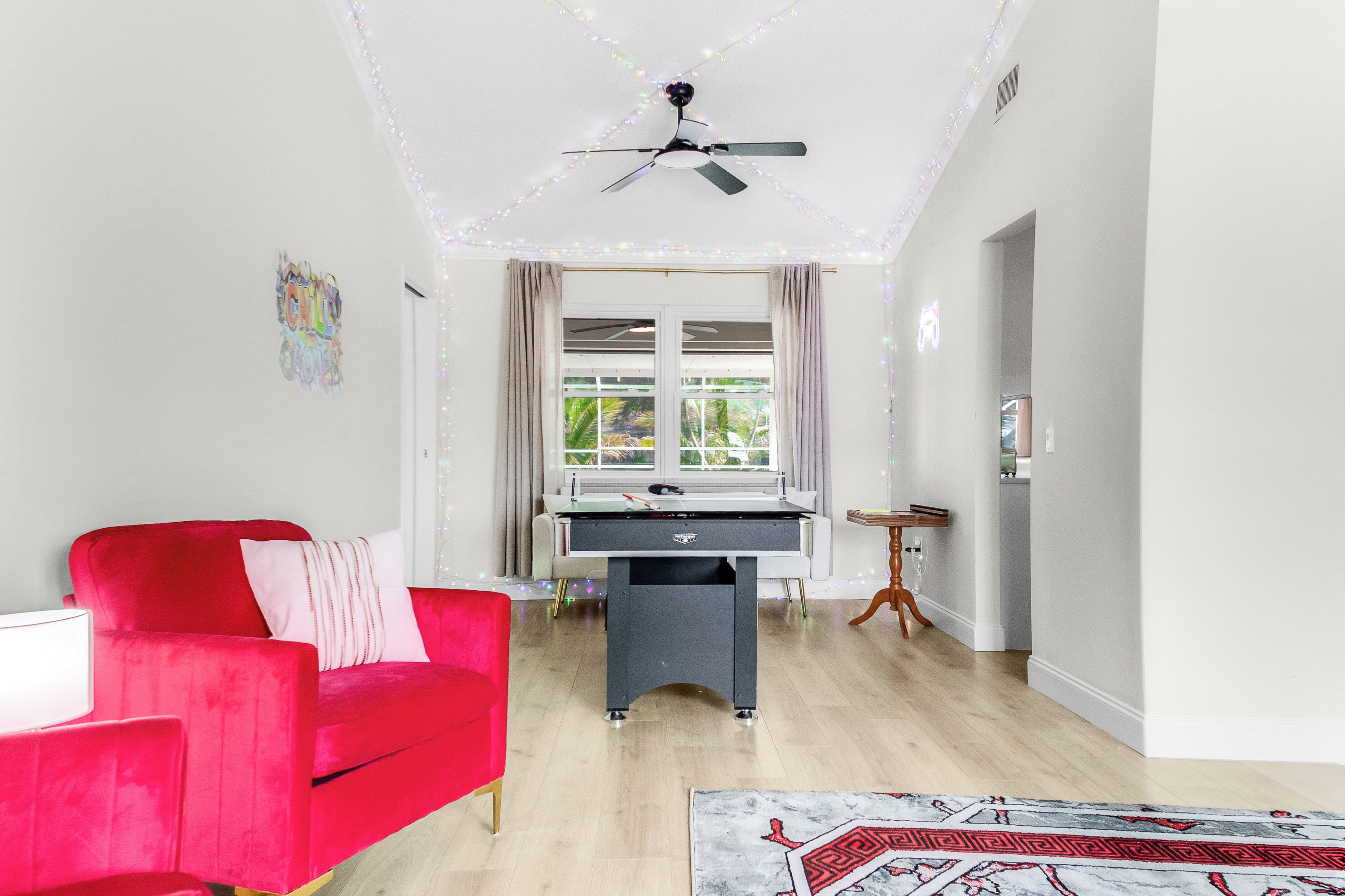 A room with a red sofa, a table with a foosball table, a ceiling fan, and a window with curtains.