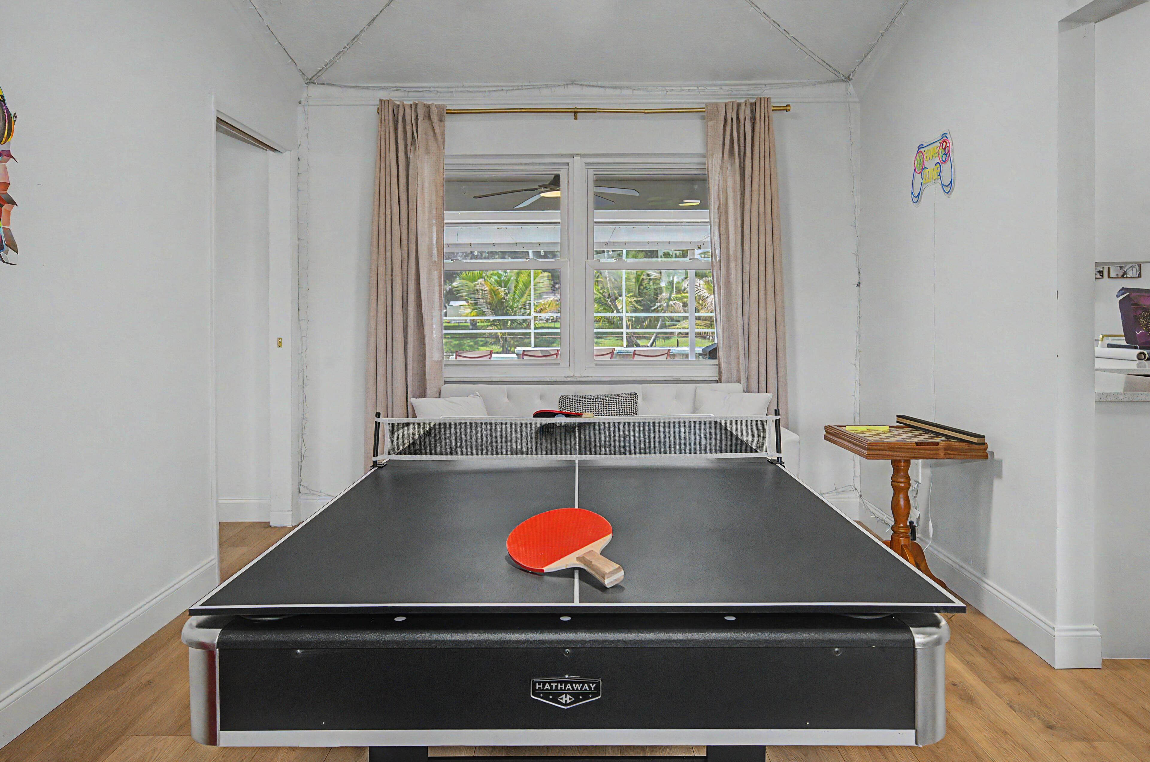 Een tafeltennistafel met een batje, een bank, een raam met gordijnen en een kleine tafel met een spel.
