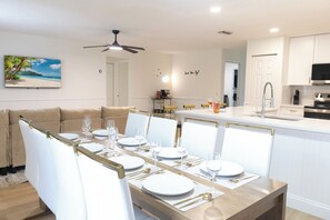 Un comedor con una mesa larga dispuesta para seis personas, una cocina con armarios blancos y un ventilador de techo.