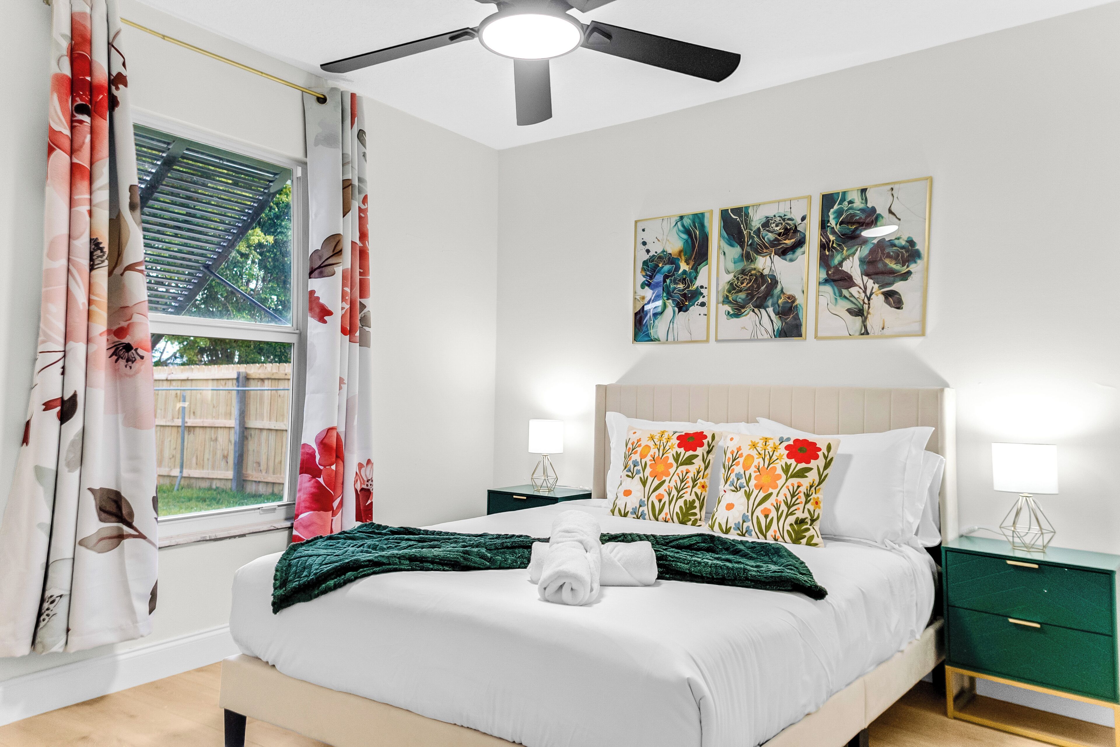 A bedroom with a bed, a ceiling fan, a window with curtains, a nightstand, and wall art.