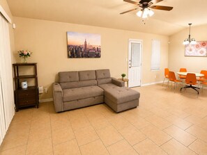 A living room with a sectional sofa, a dining table with chairs, a ceiling fan, and a cityscape painting on the wall. - 3BR Near Fort Cavazos | TDY • PCS • Extended Stay (Killeen)