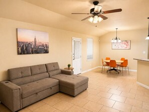 A living room with a sectional sofa, a dining area with orange chairs, a ceiling fan, and a cityscape painting on the wall. - 3BR Near Fort Cavazos | TDY • PCS • Extended Stay (Killeen)
