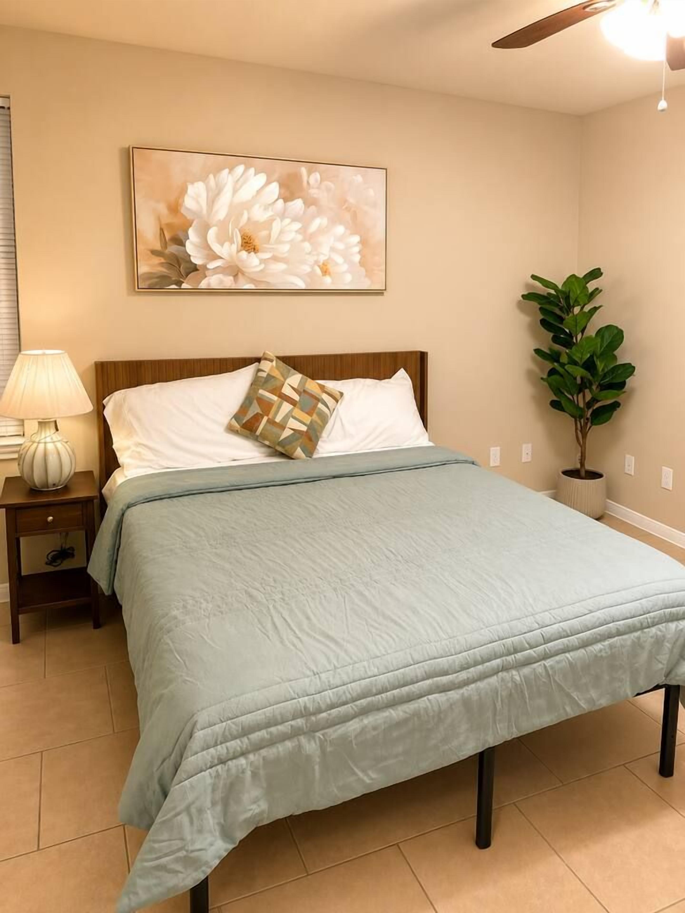 A bedroom with a bed, a nightstand, a lamp, a wall-mounted painting, and a potted plant.