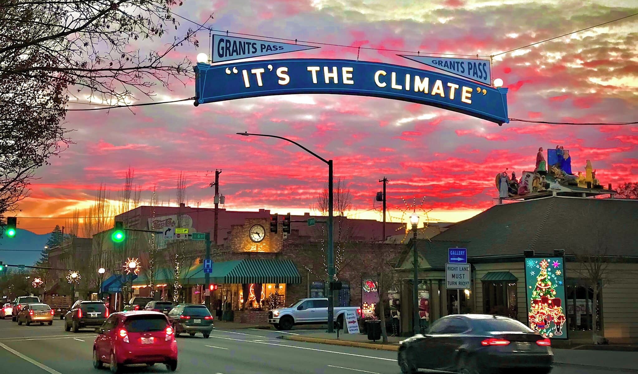 Escena callejera con un letrero destacado que dice 'IT'S THE CLIMATE' y 'GRANTS PASS'.
