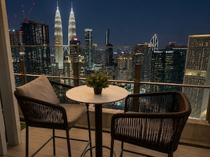 En balkon med udsigt over byens skyline, inklusive Petronas Towers, samt to fletstole med et lille bord og en potteplante.