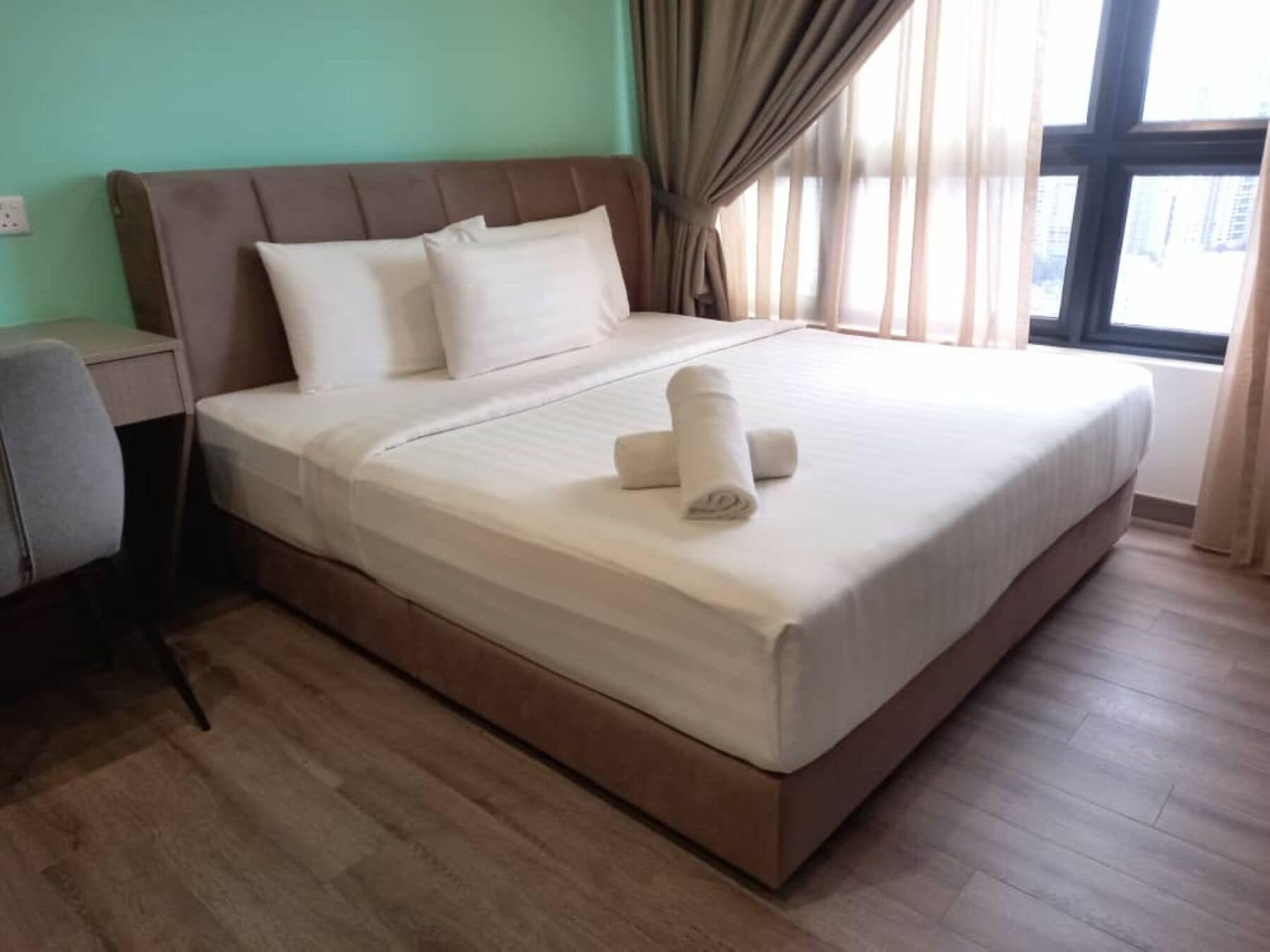 Cama bem arrumada com travesseiros e toalha enrolada, mesa de cabeceira e uma cadeira em quarto com janela grande e cortinas.