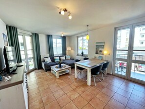 Ein modernes Wohnzimmer mit Sofa, Couchtisch, Esstisch und Stühlen. Große Fenster mit Vorhängen, eine Lampe und ein Gemälde an der Wand.