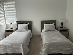 A double bed with a grey headboard, a bedside table with a lamp, and a window with blinds. - Modern Bremerton Escape Near Ferry & Seattle (Bremerton)