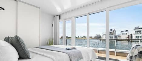 Une chambre moderne dotée d’une grande fenêtre donnant sur un plan d’eau et des bâtiments.