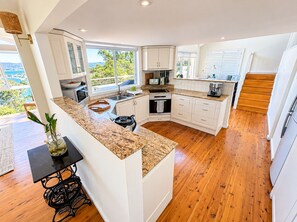 A modern kitchen with a central island, white cabinetry, and a view of the outdoors. - Carinya Coastal Escapes | Bay Views & Bouddi (Daleys Point)