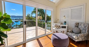 Varanda com vista para um corpo de água e colinas, uma cadeira, uma pequena mesa com abajur e um vaso de planta.