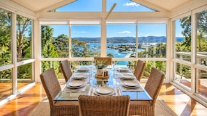 A dining area with a table set for six, surrounded by wicker chairs, large windows offering a view of a coastal landscape. - Carinya Coastal Escapes | Bay Views & Bouddi (Daleys Point)