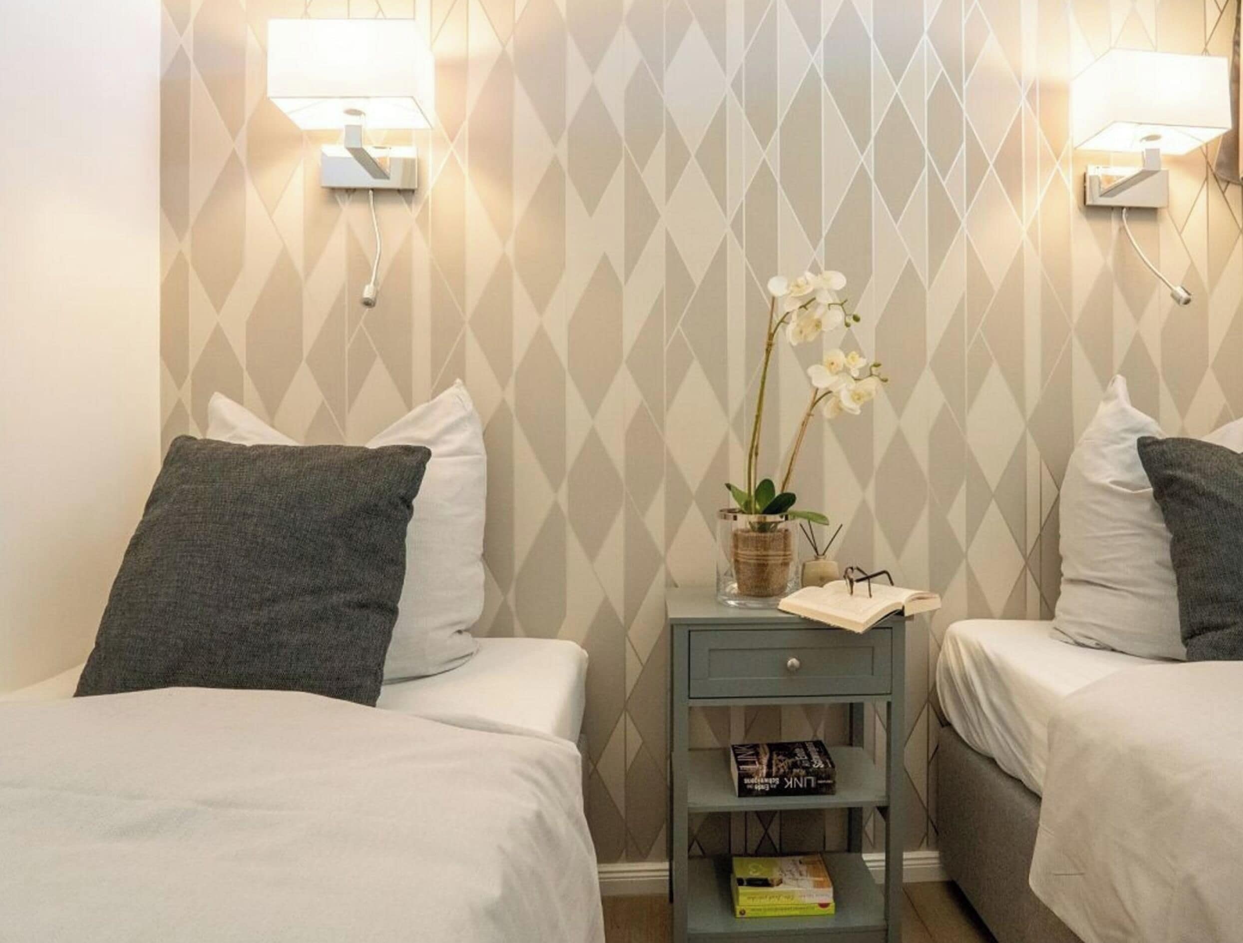 A hotel room with two beds, a nightstand with a lamp, a small shelf with books, and a decorative orchid plant.