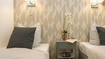A hotel room with two beds, a nightstand with a lamp, a small shelf with books, and a decorative orchid plant.