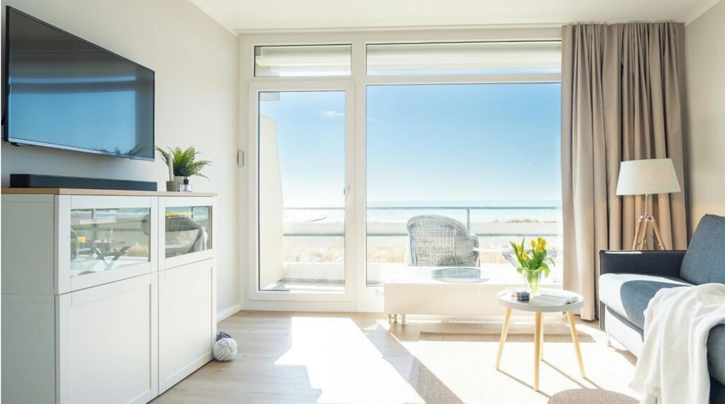 A modern hotel room with a flat-screen TV, a sofa, a small table, and a view of the sea.