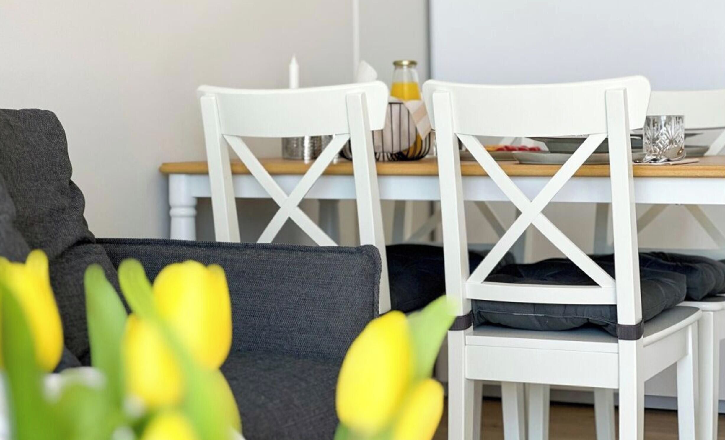 Una habitación con una mesa de comedor blanca, sillas blancas, un sofá gris y una pequeña planta con flores amarillas.