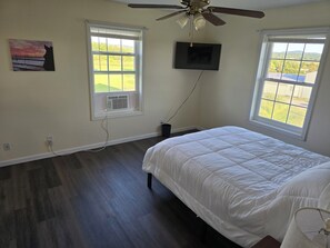 A bedroom with a bed, a ceiling fan, a TV mounted on the wall, and two windows.