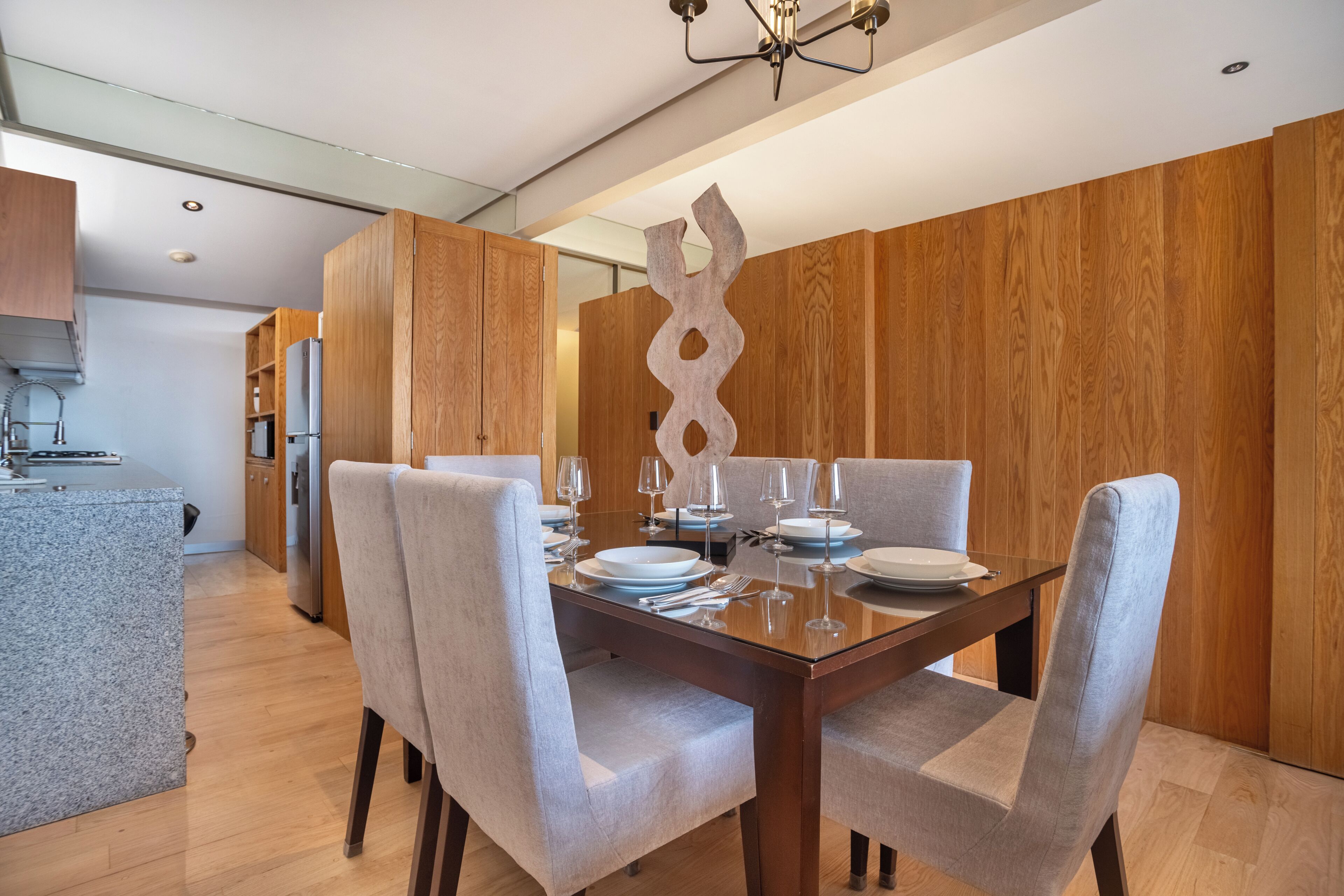 Un espace repas moderne avec une table et des chaises en bois, une cuisine en arrière-plan et un élément décoratif mural.