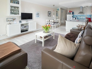 Ein modernes Wohnzimmer mit einer Couch, einem Couchtisch und einem Fernseher. Im Hintergrund befindet sich ein Küchenbereich mit einer Mikrowelle und einem Spülbecken.