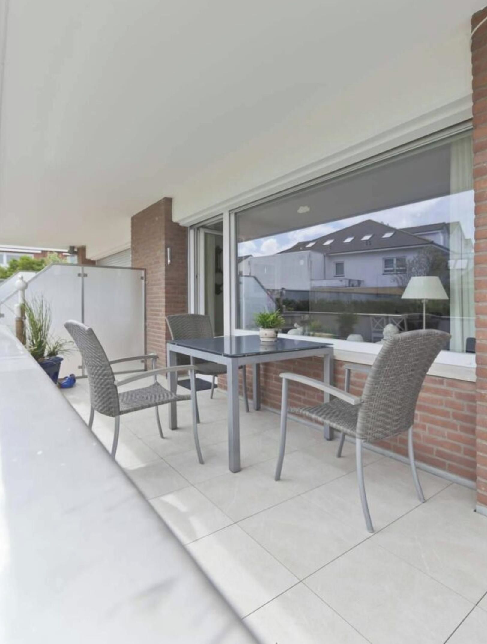 An outdoor patio with a table and chairs, a potted plant, and a view of a building through a window.