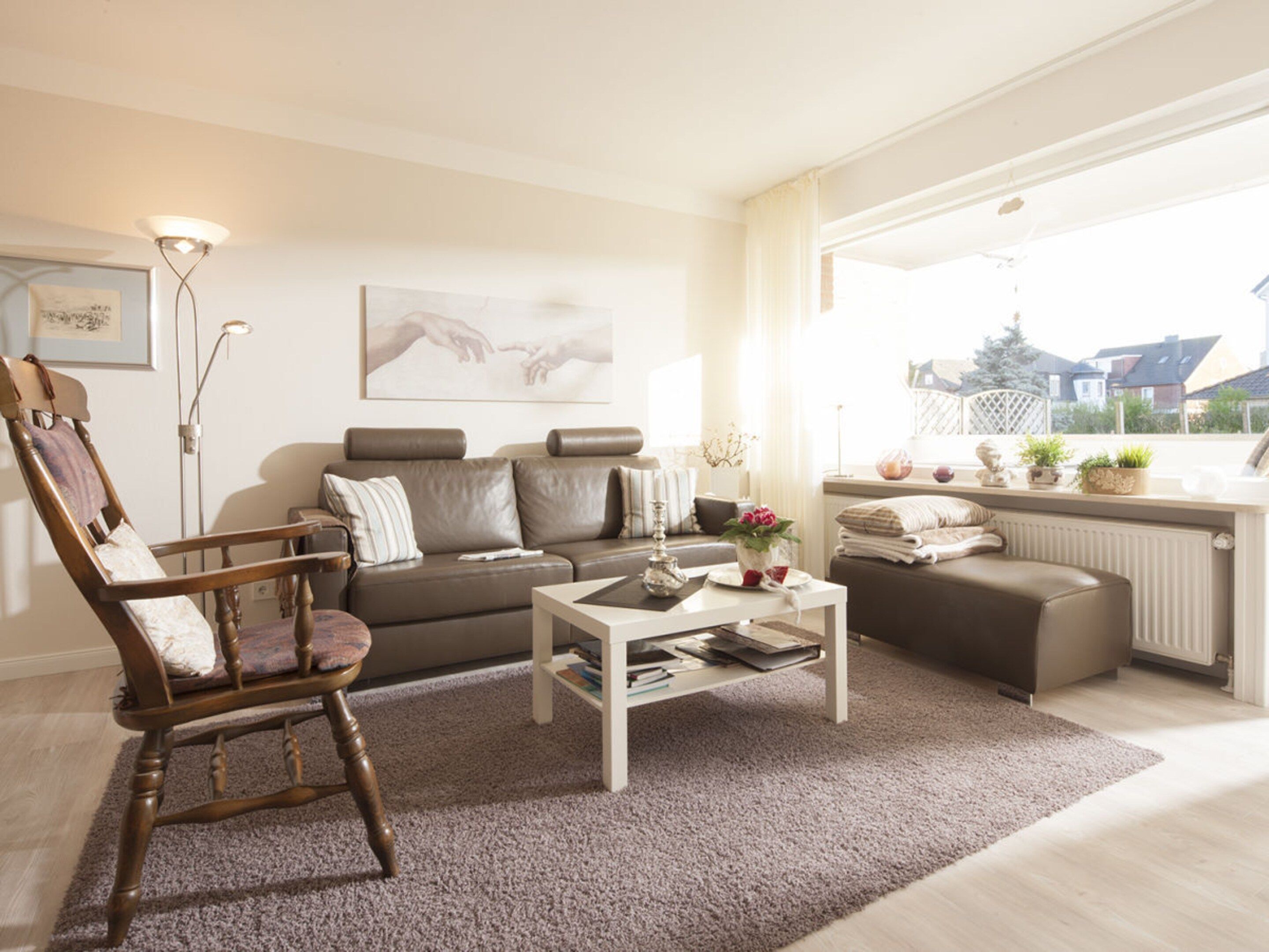 A living room with a brown leather sofa, a wooden armchair, a white coffee table, and a view of houses through large windows.