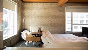 A room with a brick wall, a bed with pillows, a desk with a chair, and a window. - The Foundry Boutique Hotel (Toronto)