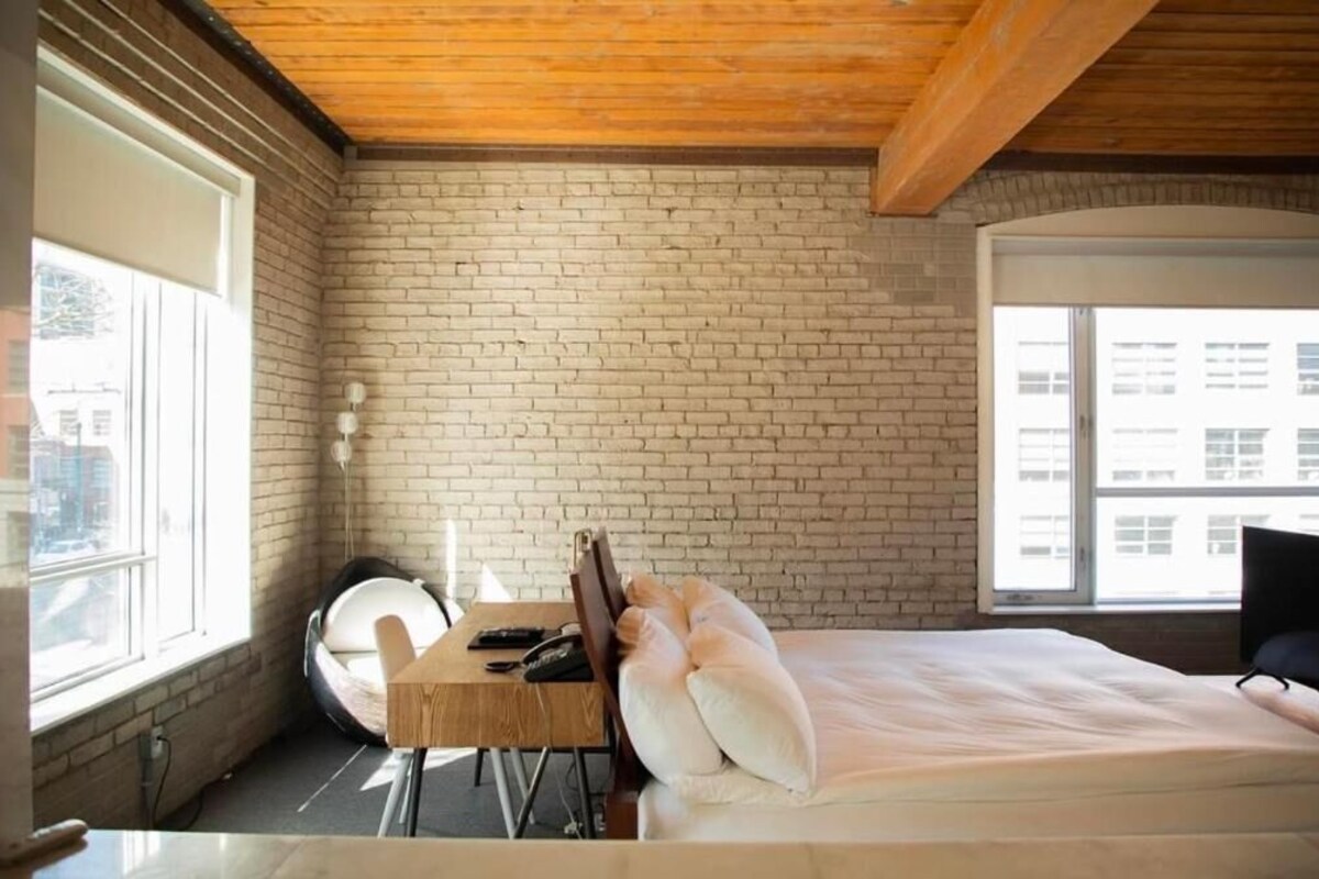 Een kamer met een bakstenen muur, een bed met kussens, een bureau met stoel, en een raam.