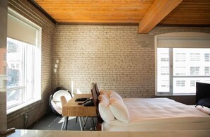 Een kamer met een bakstenen muur, een bed met kussens, een bureau met stoel, en een raam.