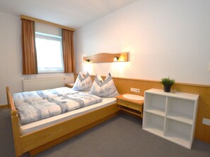 Une chambre avec un lit simple, une table de chevet, une commode, une lampe fixée au mur et une fenêtre avec des rideaux.
