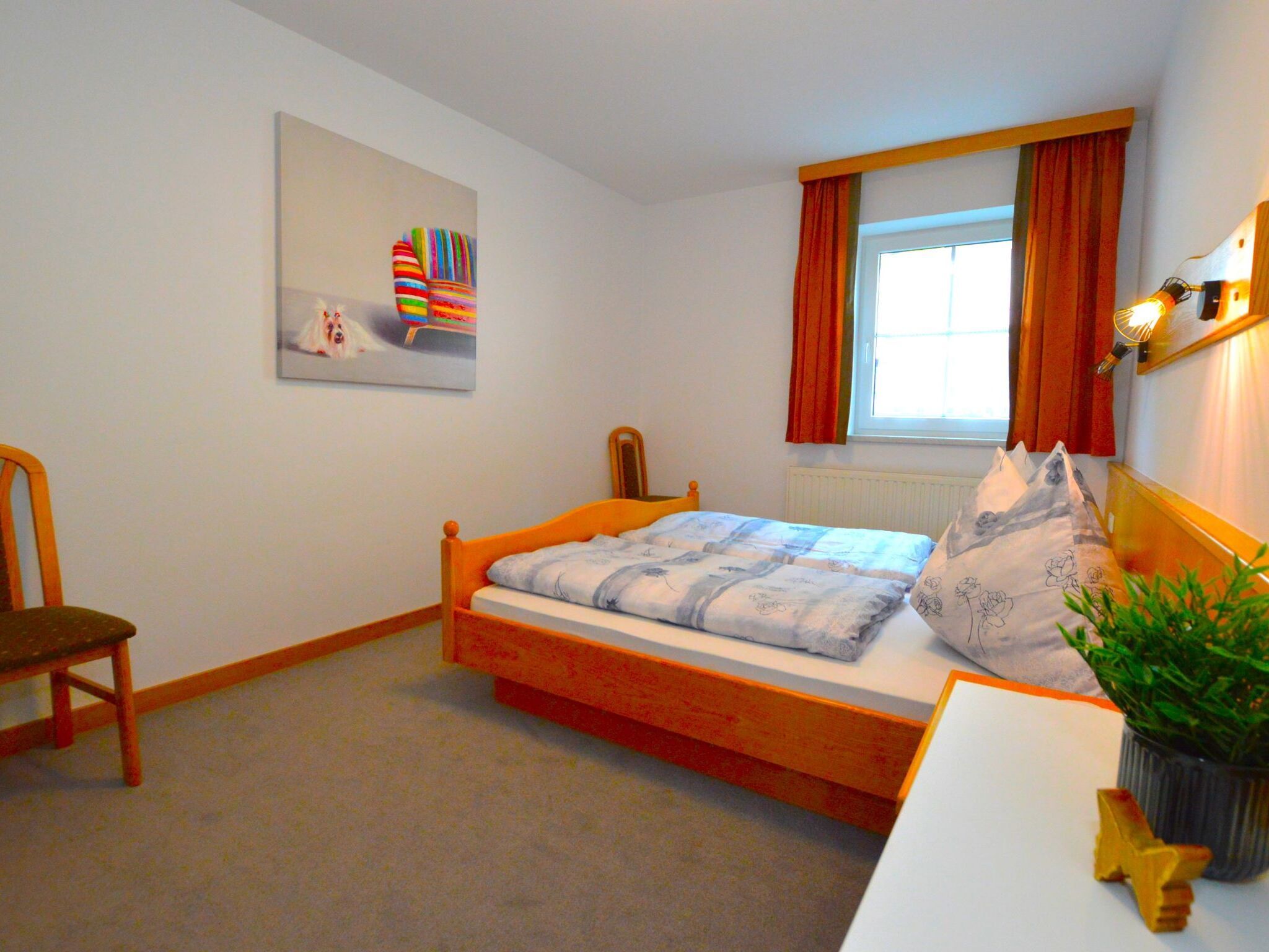 A bedroom with a single bed, a chair, a window with curtains, a radiator, and a wall-mounted lamp.