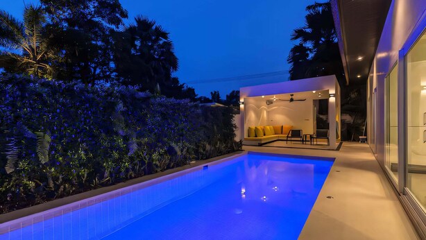 Uma área de piscina moderna com piscina azul cristalina, espaço de estar com almofadas amarelas e uma estrutura de vidro com ventoinha.