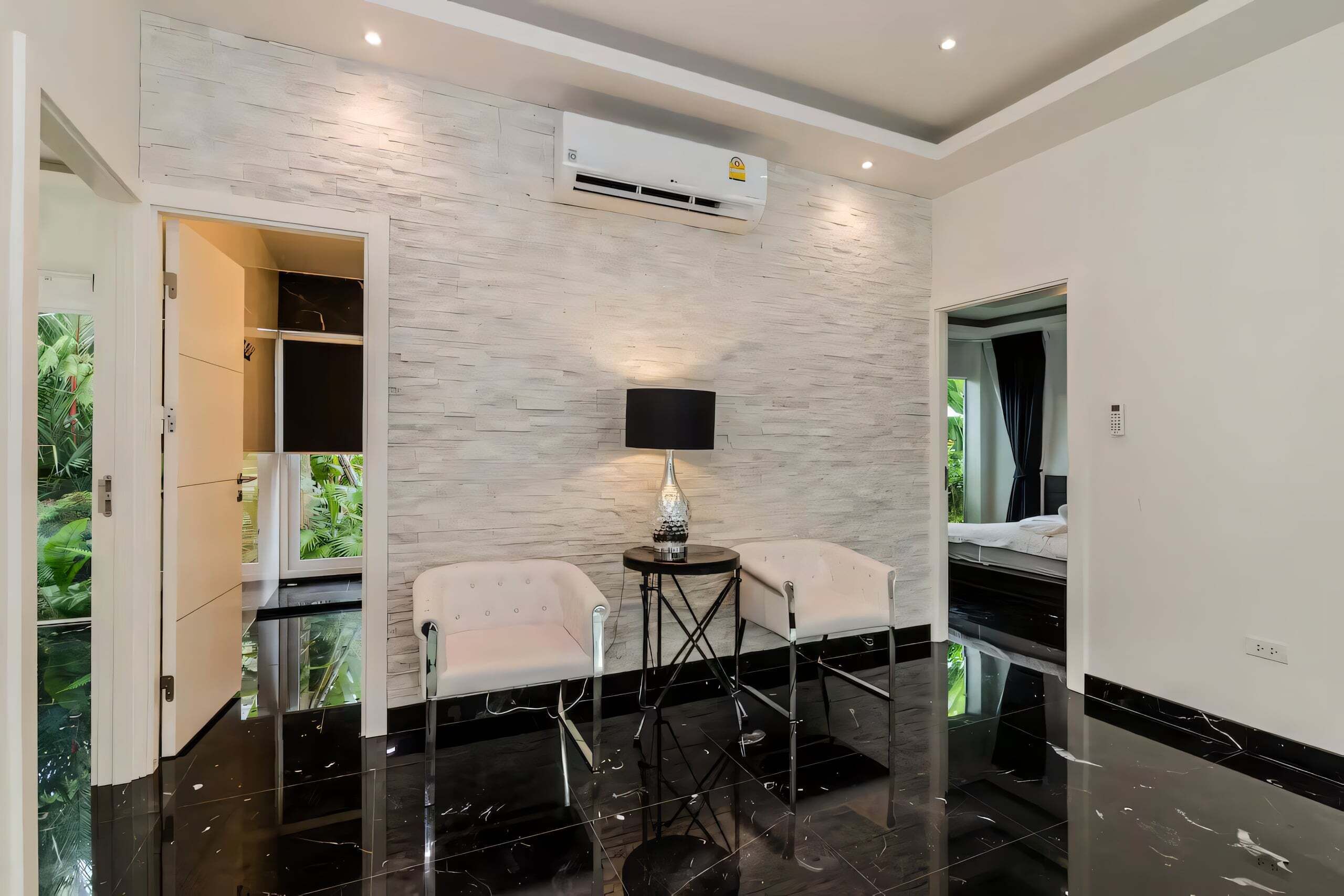 Quarto moderno com parede de pedra, ar-condicionado central, duas cadeiras, uma mesa pequena e vista para o verde através de uma porta.