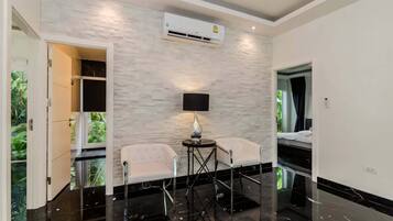 Quarto moderno com parede de pedra, ar-condicionado central, duas cadeiras, uma mesa pequena e vista para o verde através de uma porta.