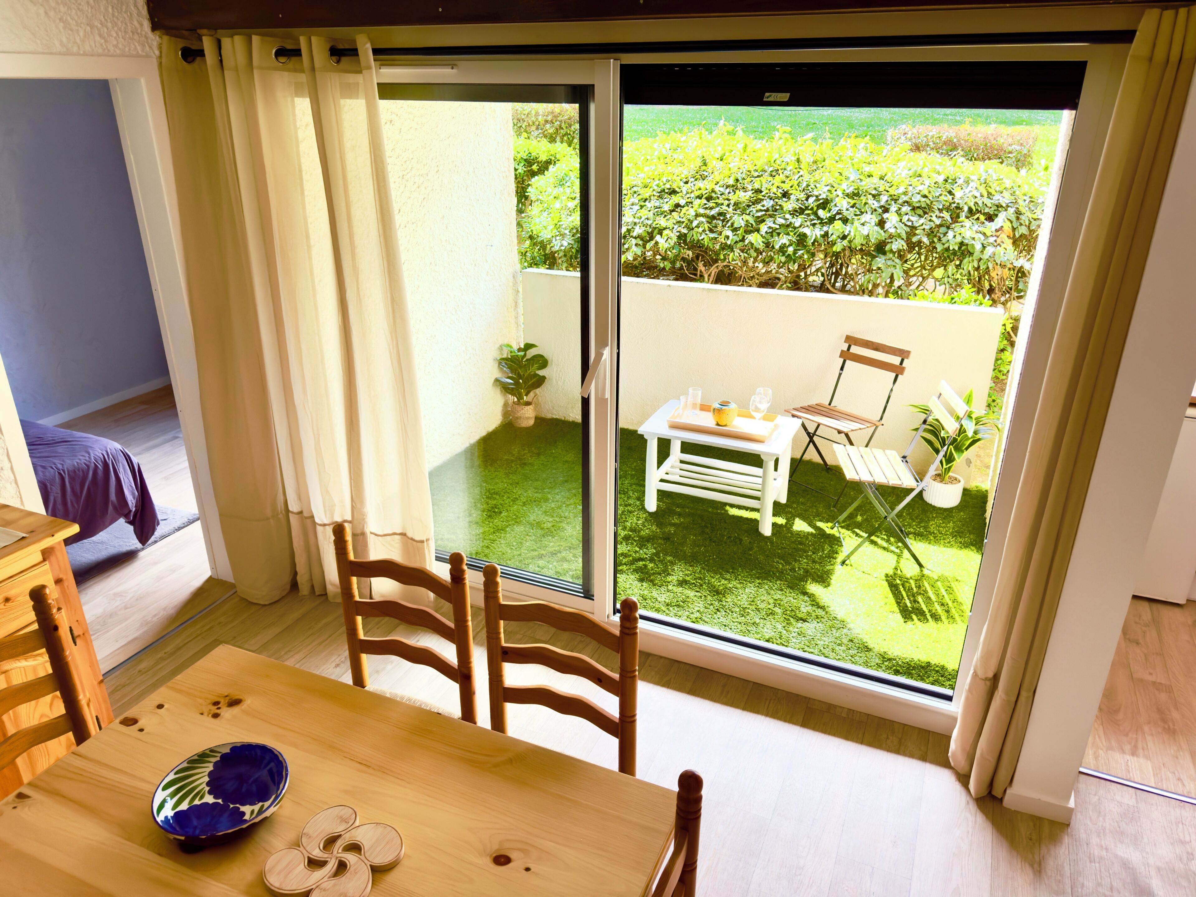 A room with a dining table, chairs, a sliding glass door leading to a balcony with a garden view.