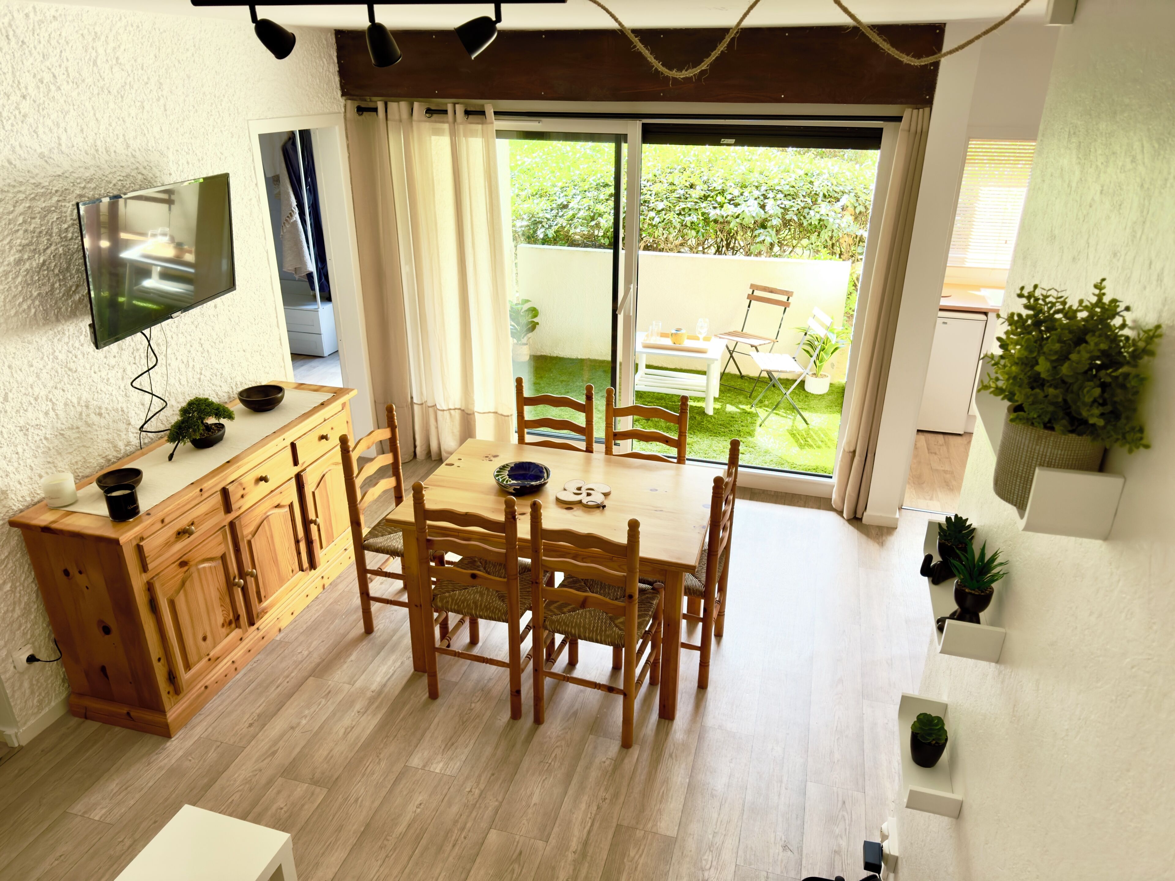 Uma sala de jantar com mesa e cadeiras de madeira, televisão fixada na parede e porta de vidro deslizante para área externa.