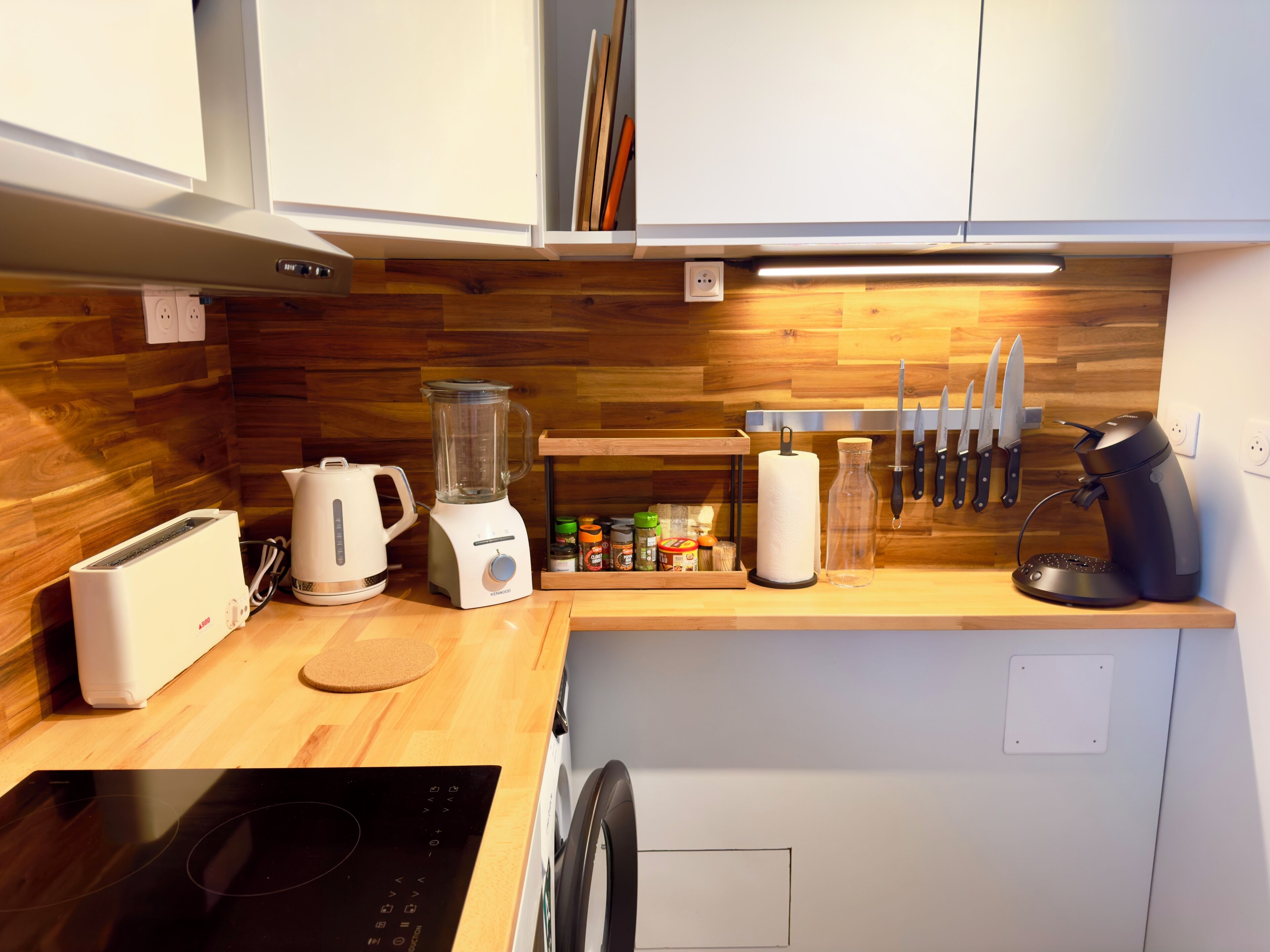 Eine moderne Küche mit weißen Schränken, einer Holz-Spritzschutzleiste, einem Toaster, einem Mixer, einem Wasserkocher, einer Kaffeemaschine und einem Messerblock.