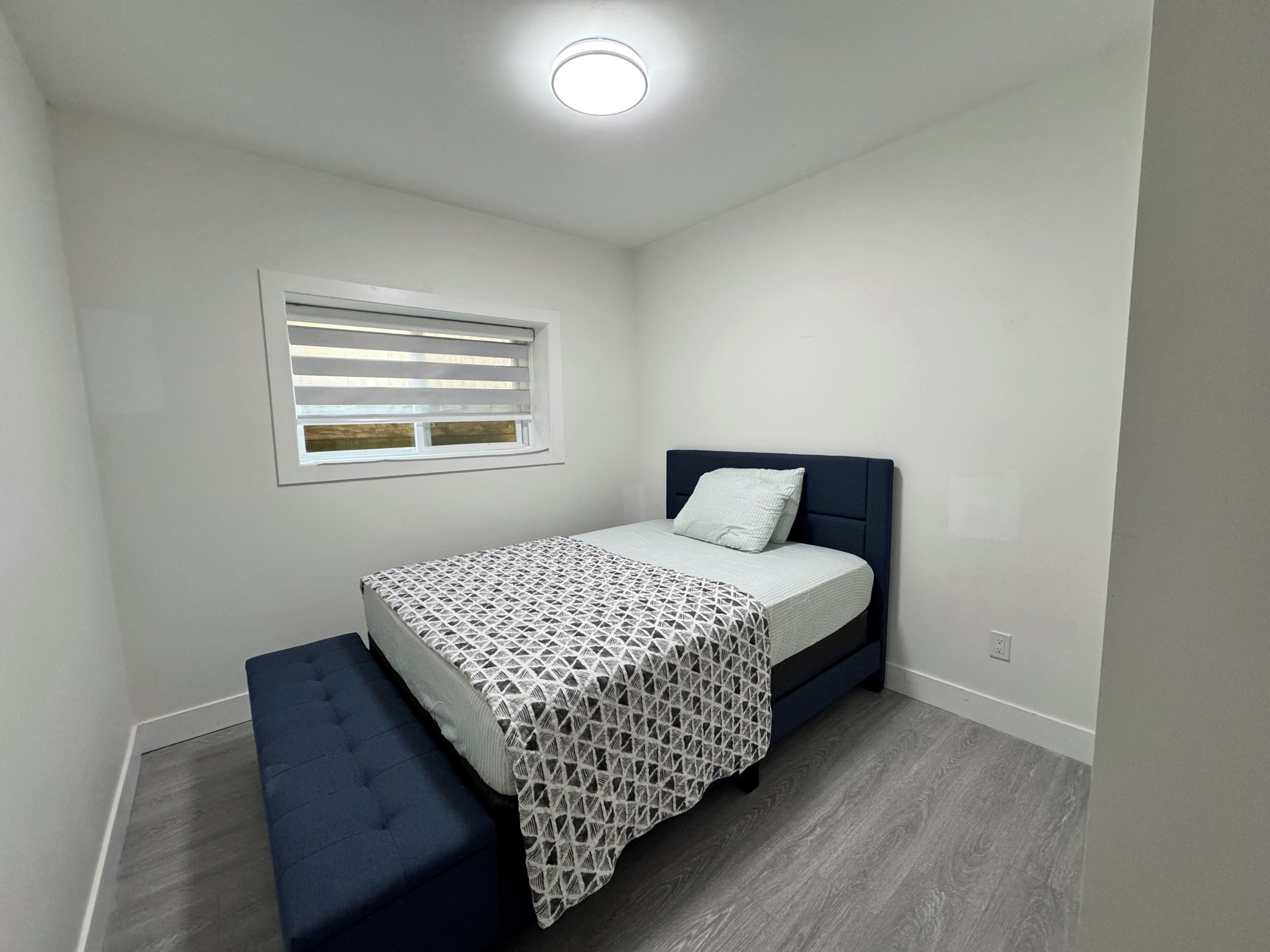 A bedroom with a bed, a bench, a window with blinds, and a ceiling light.
