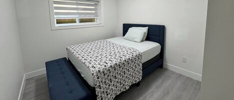 A bedroom with a bed, a bench, a window with blinds, and a ceiling light.
