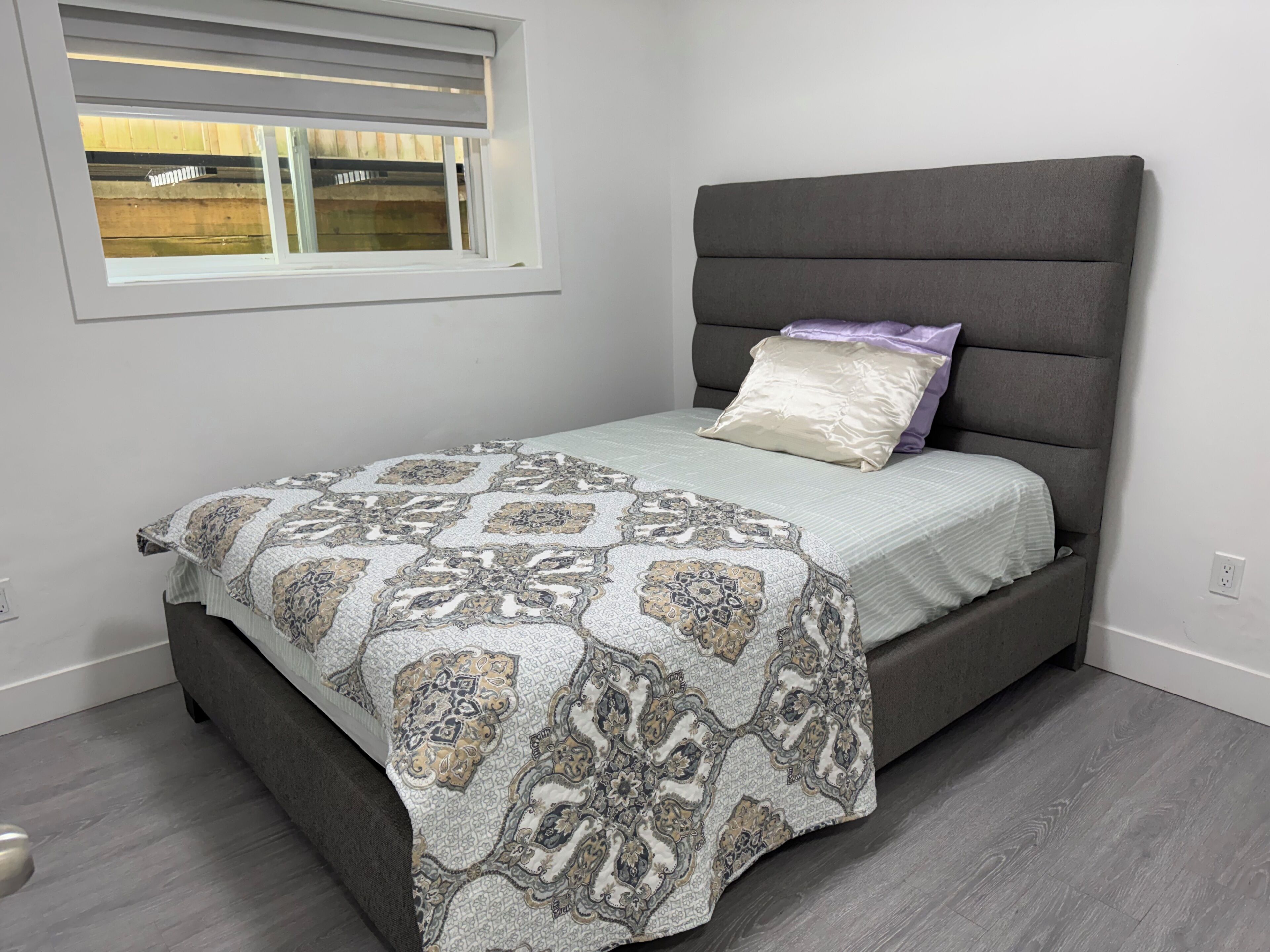 Un lit avec une couvre-lit à motifs, une tête de lit rembourrée de couleur grise et une fenêtre avec des stores.