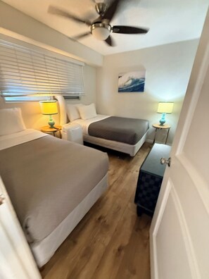 Ein Hotelzimmer mit zwei Betten, einem Deckenventilator, einem Gemälde und einem kleinen Tisch mit einer Lampe.