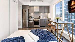 A modern hotel room with a bed, a dining table, a kitchen area, and a large window with a city view. - Eastgate Urban Suites (Toronto)