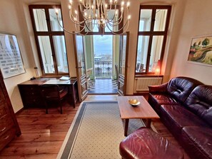 A room with a leather sofa, a wooden table, a desk with a chair, a chandelier, large windows, and a view of the outside. - Villa Bella, First row With sea View (Heringsdorf)