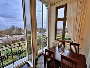 A room with a dining table and chairs, a view of a road and greenery outside, and a window with curtains. - Villa Bella, First row With sea View (Heringsdorf)