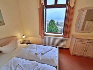 A hotel room with a bed, a nightstand, a lamp, a window with curtains, a radiator, and a wooden cabinet. - Villa Bella, First row With sea View (Heringsdorf)