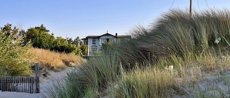 A coastal house with a sandy beach and dunes in the foreground.