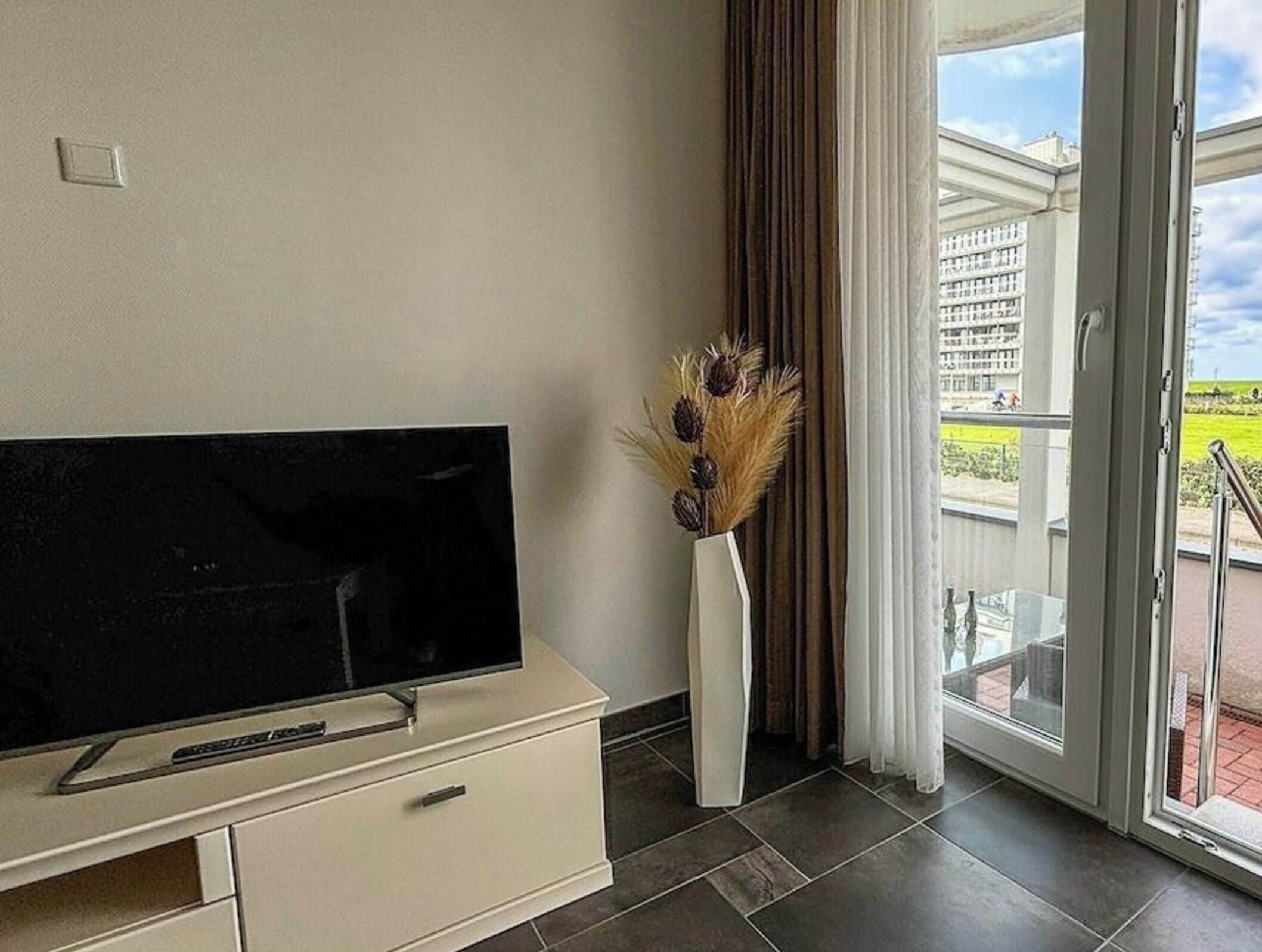 A modern living room with a flat-screen TV on a cabinet, a vase with dried plants, and a sliding glass door leading to a balcony.
